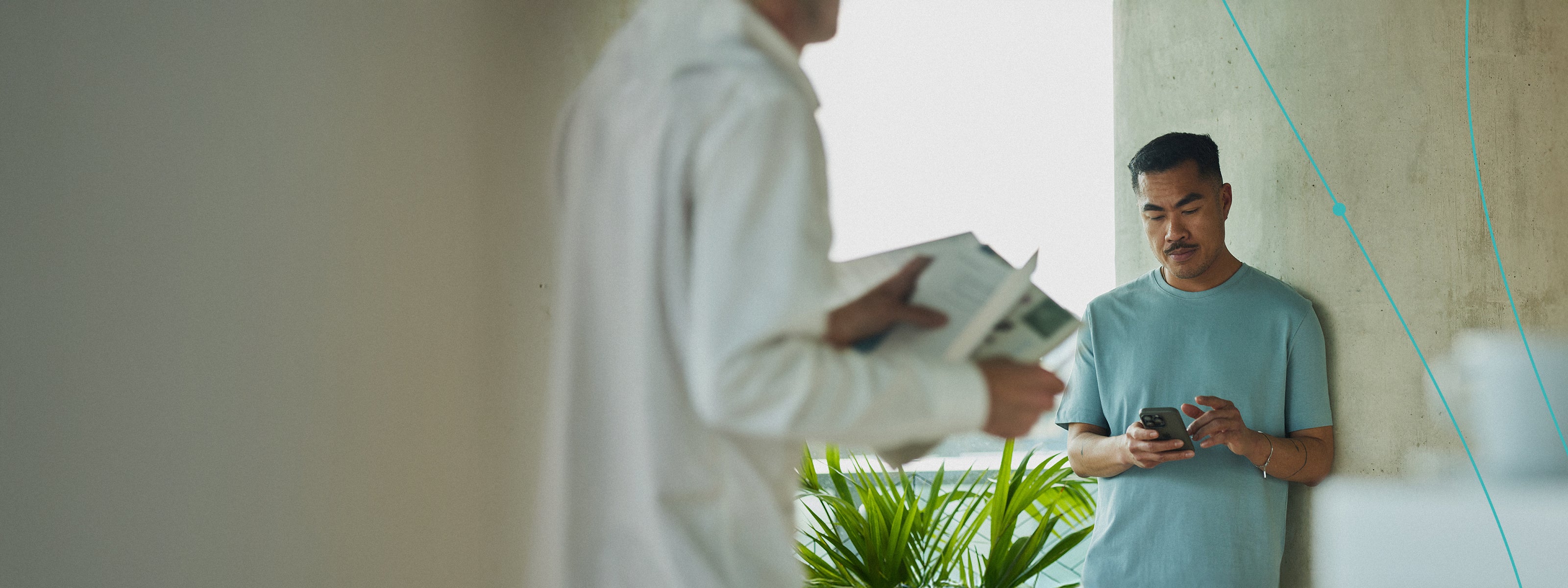 Man leaning against a wall looking at his phone, while another stands with a magazine in hand