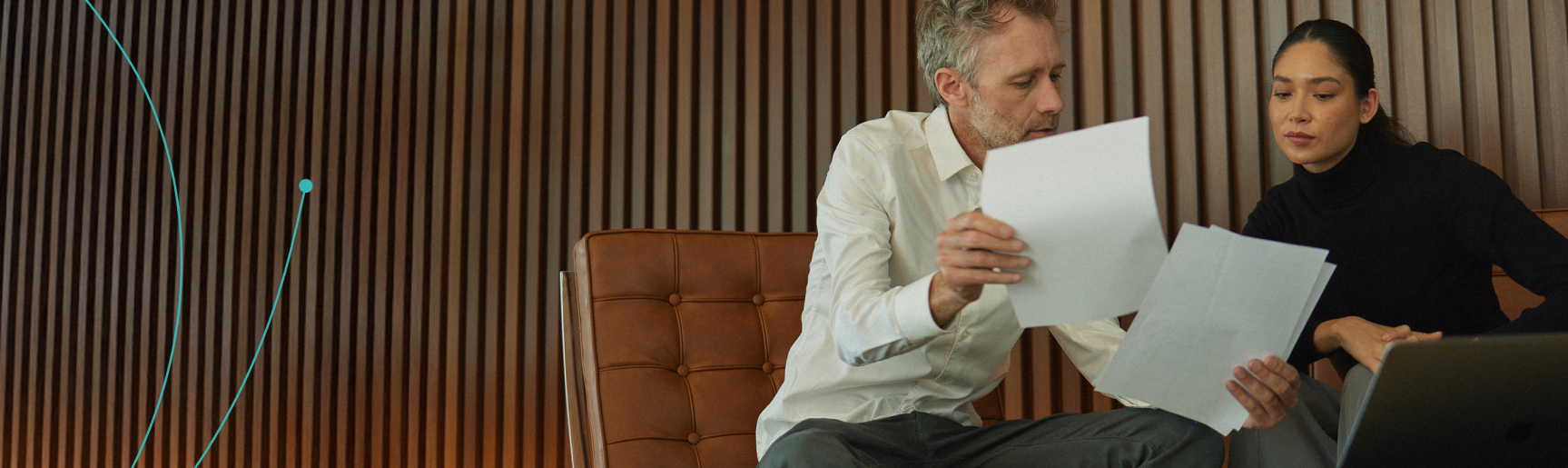 Two people sitting together, one holding papers and using a laptop, in a room with wooden paneling.