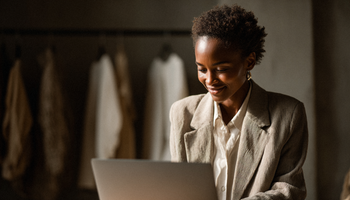 A black woman in a stylish blazer is smilingly working at her laptop. In the background you see a rack with clothes like in a showroom.
