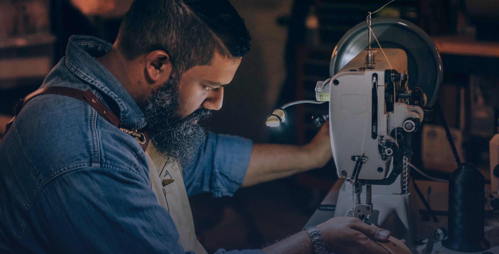 Bearded blue collar man concentrating on pushing fabric through an industrial sewing machine with an overhead, attached mini light.