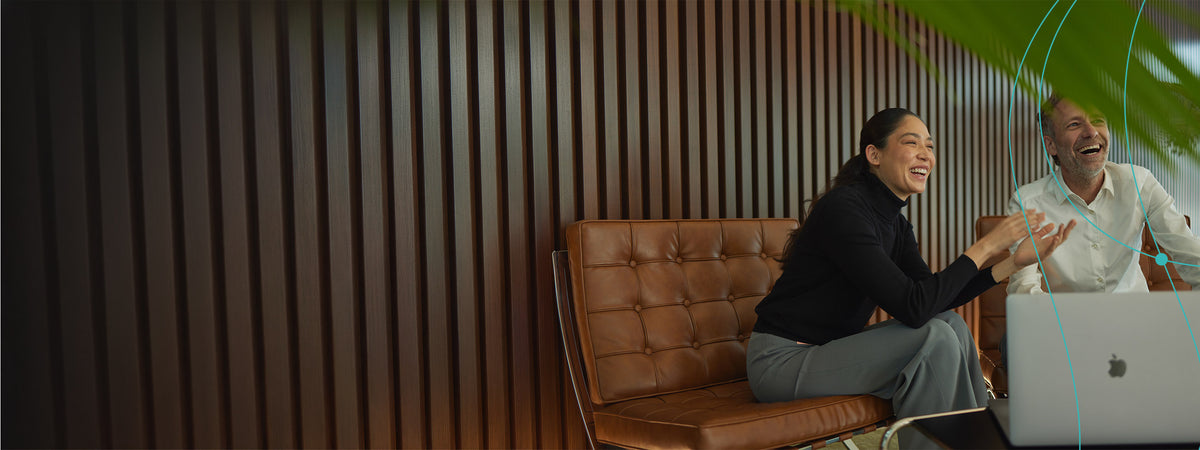 A man and a woman laughing while sitting on a brown leather bench next to a laptop in a modern office setting with a dark wood slatted wall.