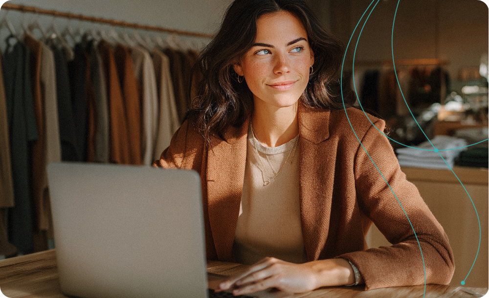 A woman with dark hair sits at a wooden desk using a laptop in a warm, softly lit boutique or office space. She is wearing a brown blazer over a beige top. - AI generated